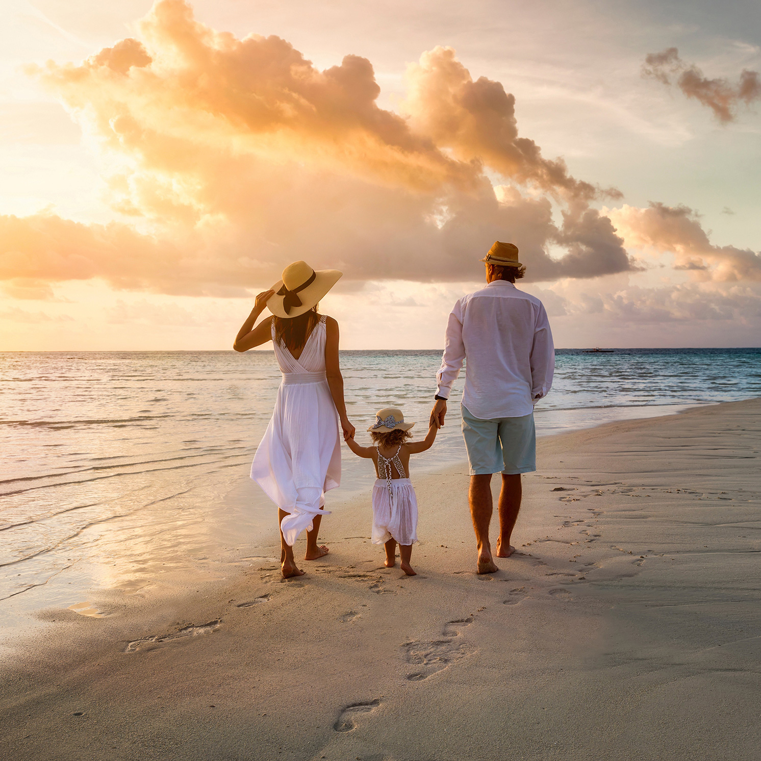 Eine junge Familie mit kleinem Mädchen geht händchenhaltend mit dem Rücken zur Kamera bei Sonnenaufgang am Strand spazieren